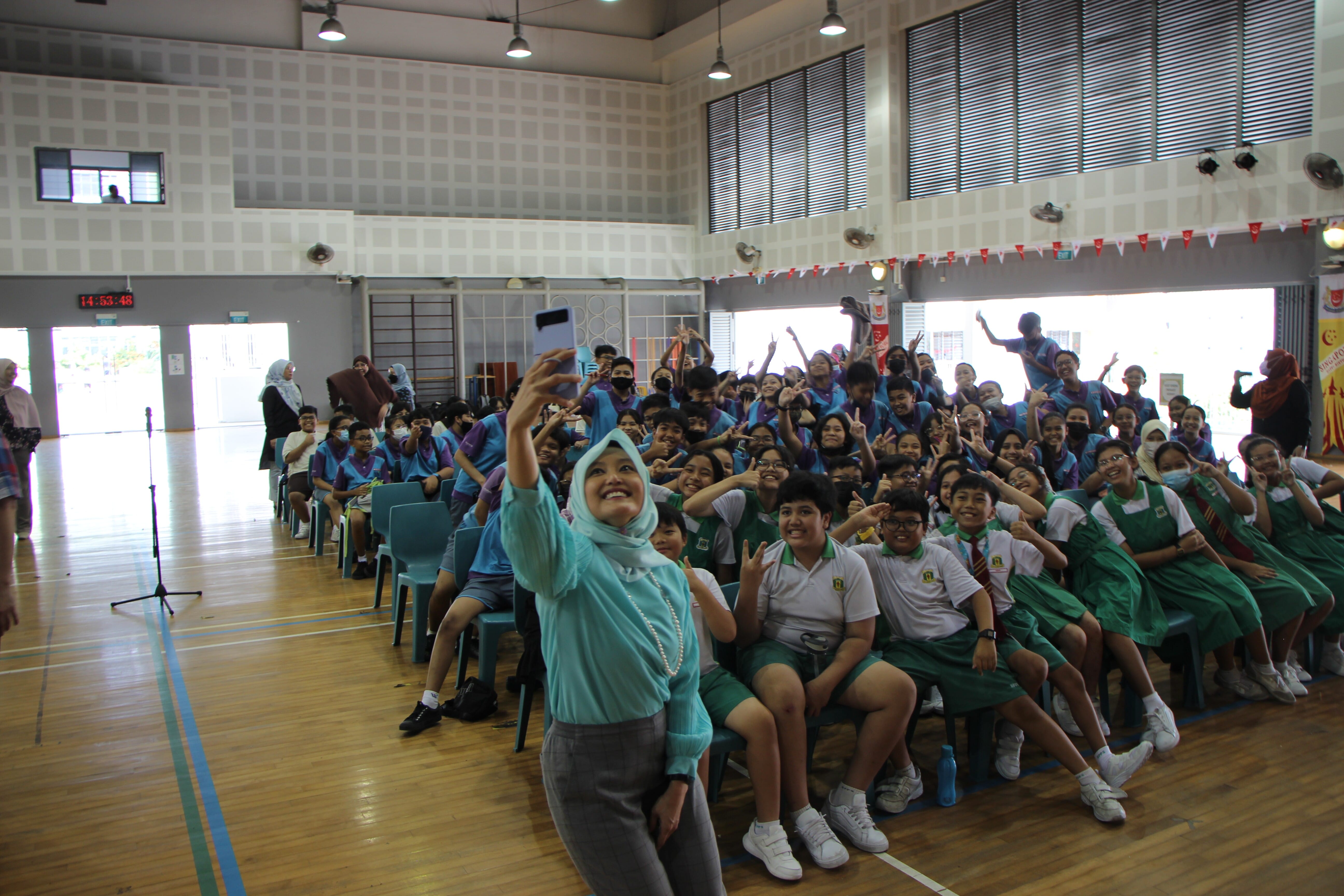 A woman in a teal hijab takes a selfie with a large group of uniformed students in a school hall.